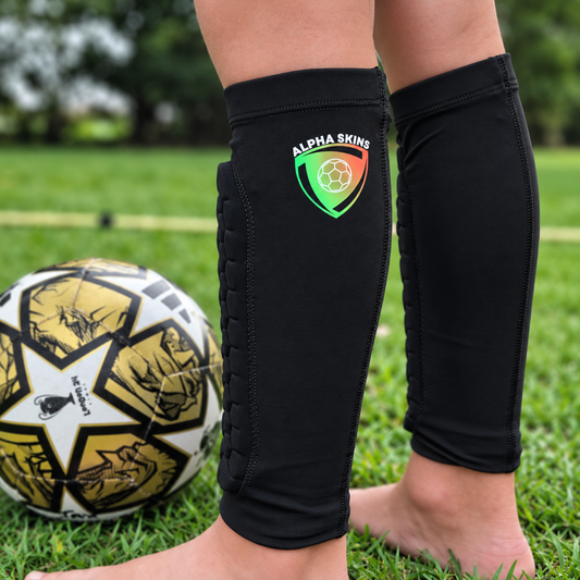 Black shin guards worn on both legs, with the left shin guard showing ALPHA SKINS logo. A soccer ball is visible on the grass next to the person's feet in an outdoor setting.