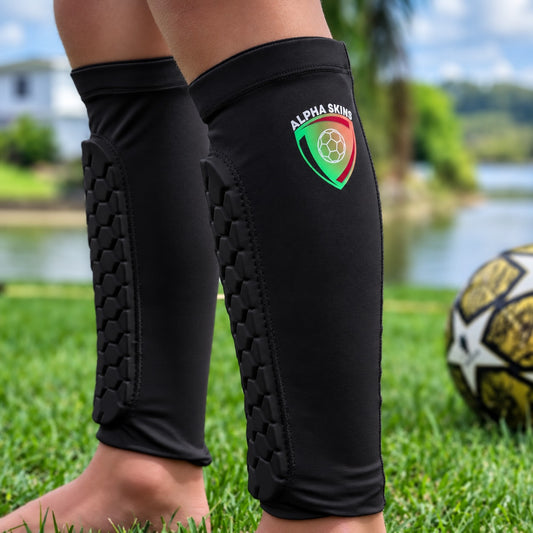 Black kids shin guards and a green, red, and white 'Alpha Skins' shield logo, worn on a childs legs on a grassy lawn with a soccer ball in the background.